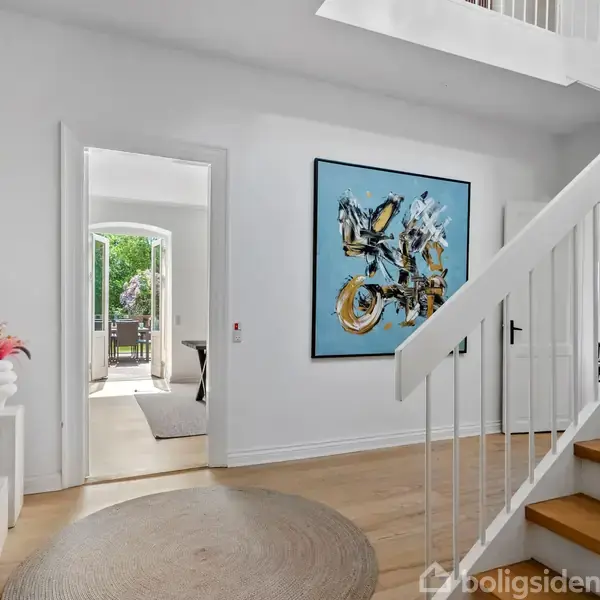 En trappeopgang med hvide vægge og lyse trægulve. På væggen hænger et stort, farverigt billede. Til venstre ses en åben dør til et værelse med udsigt til en terrasse.