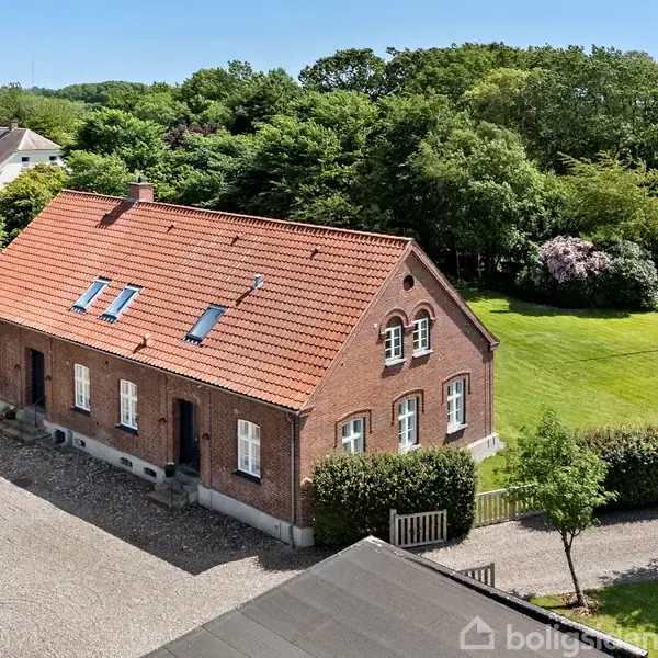 Rødt murstenshus med skrånende tegltag står i sollys. Omkranset af græsplæne og træer, fremviser det en traditionel arkitektur med flere vinduer. Grusvej og anden ejendom er synlige i nærheden.