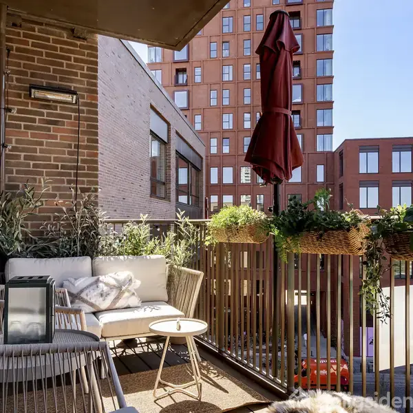 En balkon med hvide møbler og planter langs rækværket. En rød parasol er foldet sammen. Bagggrunden viser moderne, høje bygninger. Skilt med teksten "Tils" ses i gården.