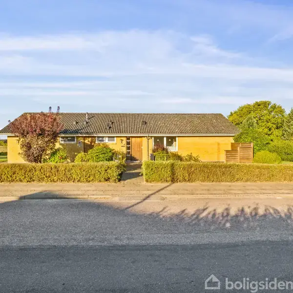 Hus med gult murværk står stille bag en lav hæk. Sollys belyser facaden, og træer omgiver området med himmel og vej i baggrunden.