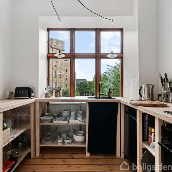 Et lille køkken med åbne hylder fyldt med glas og tallerkener. Vindue viser udsigt til en bygning og træer. Køkkenmaskiner inkluderer brødrister og elkedel på bordpladen.