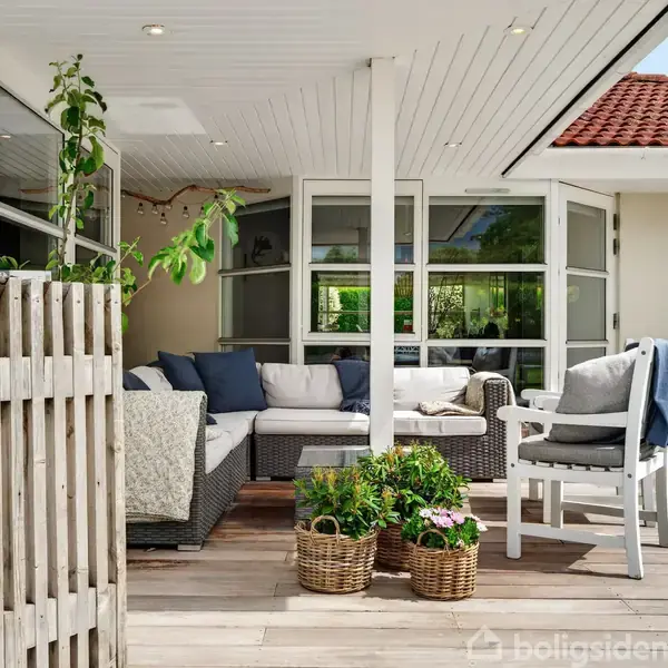 Terrasse med flettede møbler arrangeret omkring et sofabord, placeret i et hjørne med overhæng. Kurve med blomster står på plankegulvet. Ruder og åben dør i baggrunden.