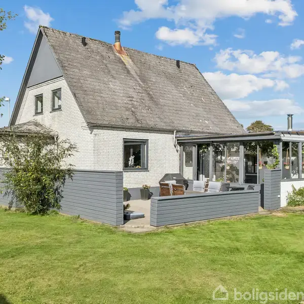 Et hvidt hus med sort tag står omgivet af grønne græsplæner. Terrassen foran har møbler til afslapning. Blå himmel og spredte skyer danner baggrunden.