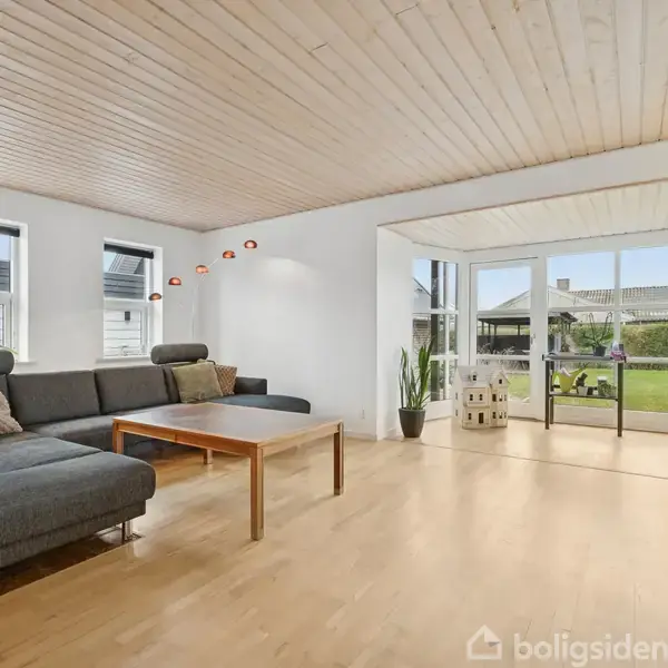 Trappe med trætrin fører op, mens lang spisebord med klar dug og moderne stole står ved siden af. Store vinduer og åbne døre tillader lysindfald i rummet.