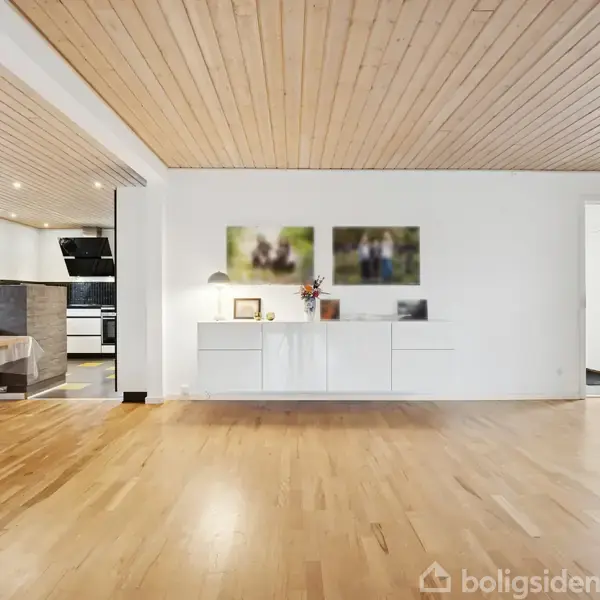 Sort hjørnesofa står i en lys stue med trægulv, flankeret af et træbord. Loftslamper hænger over sofaen, og to små vinduer med udsigt er i baggrunden.