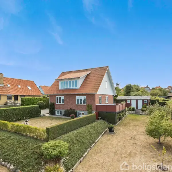 Et rødt murstenshus med trekantet tag står i en velplejet, indhegnet have med klippede buske. Det er omgivet af andre huse under en klar blå himmel.