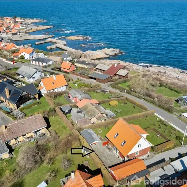 Hus med rødt tag vises fra luften, beliggende nær en kystby med flere andre huse og en vej ned til havet. Kysten strækker sig langs den klare blå havflade.