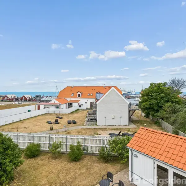 Et hus med orange tag står i en have med græs og buske. Terrassen har møbler, og et hvidt hegn omgiver området. Havet kan ses i baggrunden under en næsten skyfri himmel.