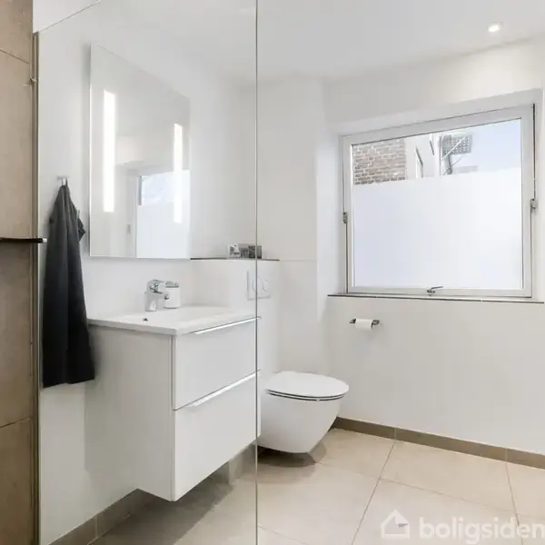 Badeværelse med bruser til venstre, hvid håndvask og spejl i midten, toilet ved vindue til højre og vaskemaskine yderst til højre.
