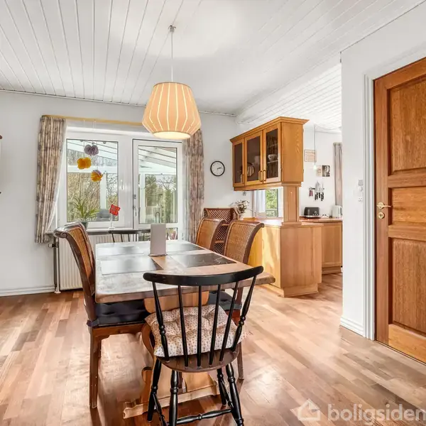 Langt spisebord med stole i en lys spisestue; store vinduer tilbyder udsigt til haven. Træskabe og en dør er i nærheden. En hængelampe lyser rummet.