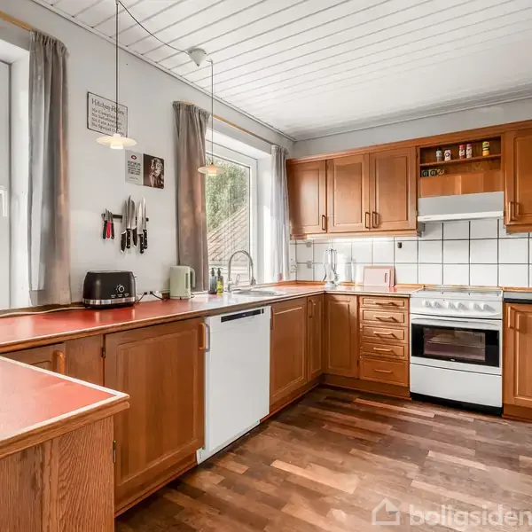 Et køkken med træskabe og hvide fliser. Bordet har blandt andet en brødrister og knive. To store vinduer giver udsigt til træer. Gulvet er af mørkt træ.