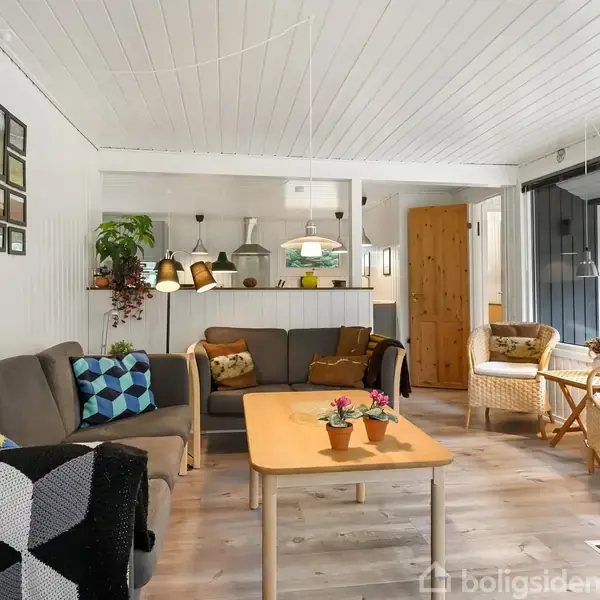 En stue med grå sofa og træbord omgivet af stole. Væggene er dekoreret med billeder. Der er grønne planter, pendellamper og store vinduer. Tæpper pryder stolene.
