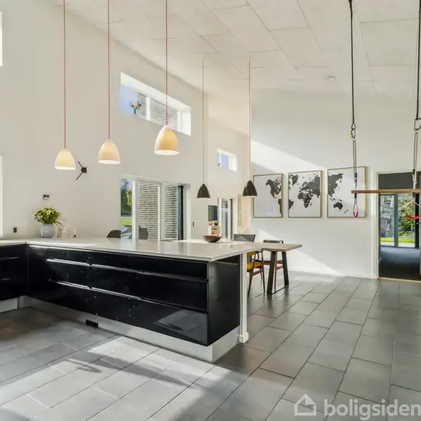 Køkken med sort L-formet køkkenø og hængende lamper. I baggrunden er der spiseplads med et bord og stole. Væggene har store vinduer og verdenskort som dekoration.