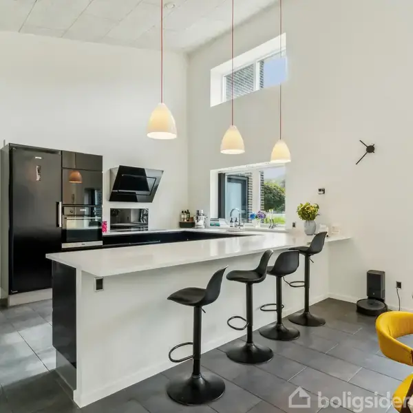 Et moderne køkken har en stor, hvid bardisk med tre sorte barstole. Hængende lamper oplyser området, mens store vinduer giver naturligt lys. Sorte apparater står langs væggen.