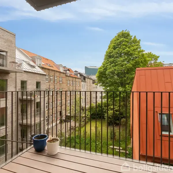 En balkon med to potteplanter står stille. Den omfatter et udsyn til flere bygninger i mursten og træer, der omgiver en grøn gård i et boligområde.