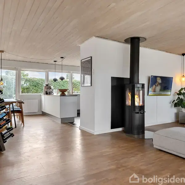 Rummeligt opholdsområde med stilfuldt møblement, åbne skydedøre til terrasse, omkranset af store vinduer. Terrasse med sofa og udsigt til grønne træer skaber en indbydende atmosfære.