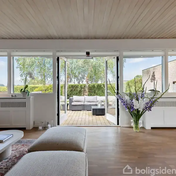 Reol fyldt med vinflasker står vinkelret på en åben stue med sofa og brændeovn. Et moderne køkken ses til venstre med store vinduer og spisebord.