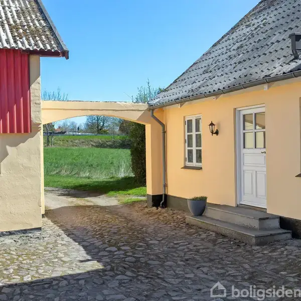Et gult hus med en hvid dør og vinduer står stille ved siden af en gårdsplads med brostensbelægning. En stilfuld bue forbinder bygningen med en rød lade.