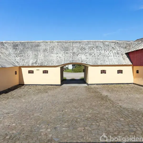 Gårdsplads omgivet af tre længer; to gule bygninger og en rød fjøs. Stenbelagt gårdsplads med åbning til en blå himmel i baggrunden.