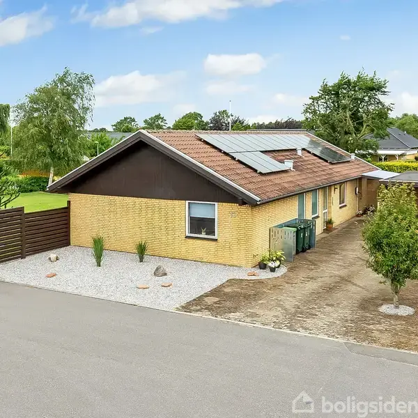 En enkeltfamiliehus i gule mursten med solpaneler på taget står ved siden af en indkørsel. Haven indeholder spredte små træer. Omgivelserne inkluderer en vej og andre huse.