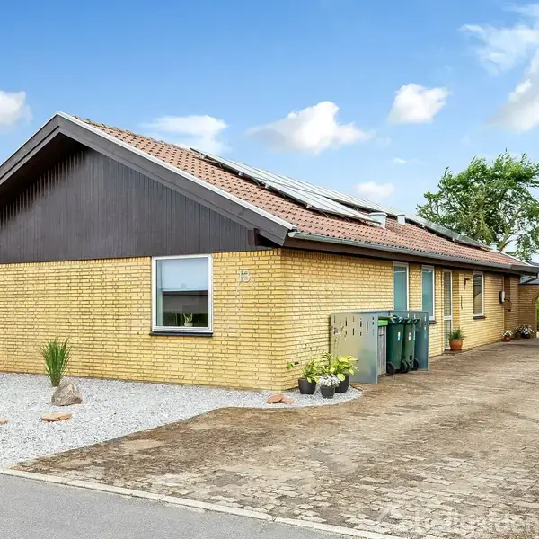 Et gult murstenshus med skråt tag og solpaneler står langs en indkørsel. Foran huset er der grus og planter. Nummer 75 ses ved vinduet.