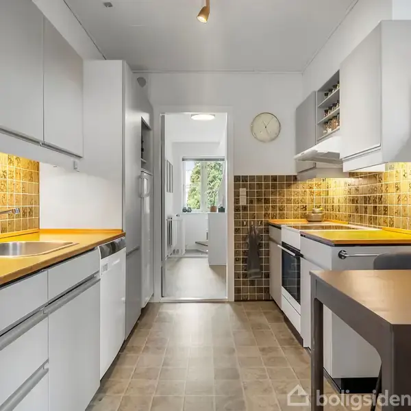 Moderne køkken med hvide skabe og gule bordplader. Kogeplade og ovn ved væggen. Mosaikfliser på væggen. Et rundt ur hænger over døren til en lys korridor.