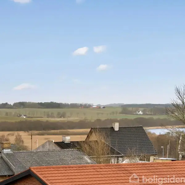Rødt tegltag ses i forgrunden med flere hustage omkring, under en klar blå himmel. I baggrunden er der åbne marker og en stor, stille sø omgivet af træer.