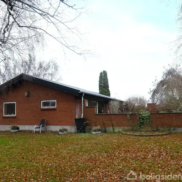 En rød murstenshus står i en have dækket af nedfaldne blade. Der står to stole ved siden af huset. Træer uden blade omgiver området.