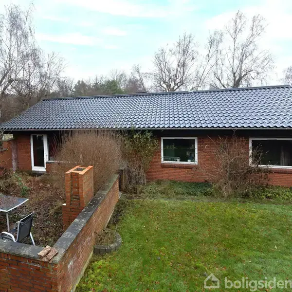 Et rødt murstenshus med sort tag står midt i en have omkranset af buske og træer. En lille muret skorsten er tilknyttet en terrasse med et bord og en stol.