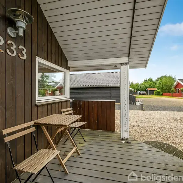 Træveranda med bord og to stole. Husnummeret "233" er synligt på væggen. Kontekst er en grusbelagt gård med udsigt til røde huse i baggrunden under en blå himmel.