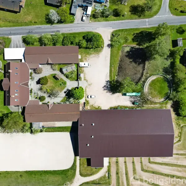 Bygning set ovenfra med flere røde tage, omkranset af grønne arealer og indkørsel. To biler holder parkeret. En større bygning ses mod bunden, og en lille runding mod højre.