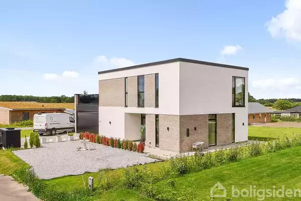 En moderne, to-etagers bolig med fladt tag står på en grusbelagt gårdsplads. Til venstre holder en varevogn parkeret. Omkring er der grønne marker og et andet hus i baggrunden.