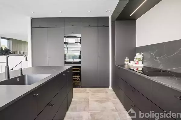 Et moderne køkken med sorte skabsfronter. En indbygget ovn og vinkøler er integreret i midten af skabene. Bordpladerne har en mørk marmoroverflade. Gulvet er lyst flisebelagt.