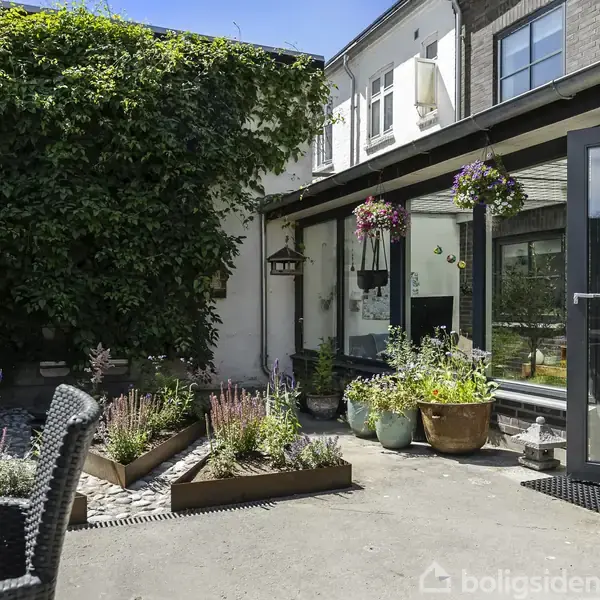 Terrasse med plantekasser og blomsterpotter, omgivet af en bygning med store vinduer. Hvide og grå vægge, dør åben mod haven. Møbler og grøn bevoksning i nærheden.