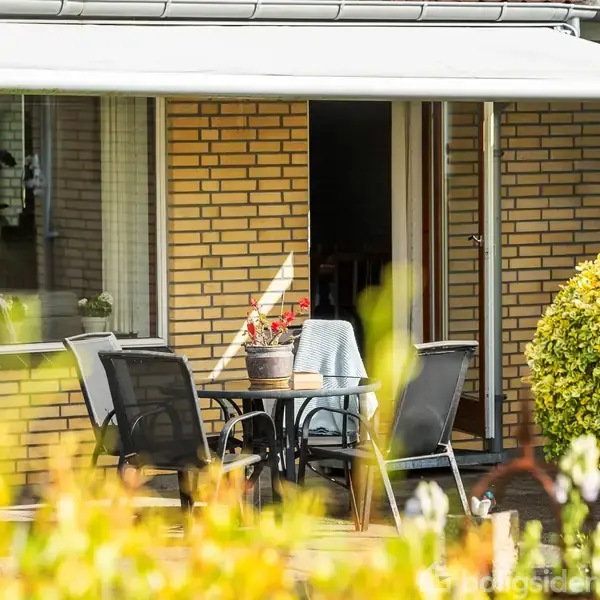 Terrassebordet står med fire stole omkring, dekoreret med en blomsterkrukke. Det hele er placeret ude foran en murstensvæg og under en markise. Buske omgiver området.