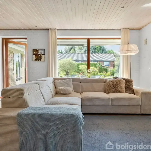 Stor lys stue med stor, lys grå hjørnesofa dækket med puder, placeret på blå tæppe. Store vinduer giver udsigt til have. Træloft, hvidmalet væg og skab til venstre.