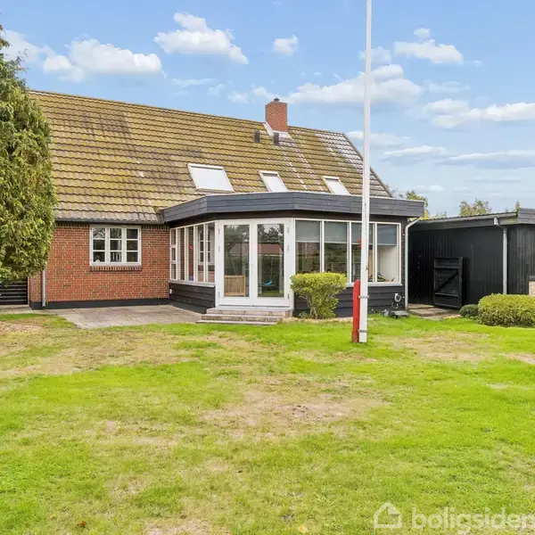En rød murstensvilla med karnap og garage, omkranset af en græsplæne. Et flagstang står i haven, og himlen er blå med spredte skyer.