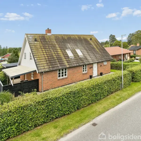 Rødt murstenshus med skråt tag og tagvinduer, omgivet af hække og andre huse i et villakvarter, under en blå himmel med skyer.