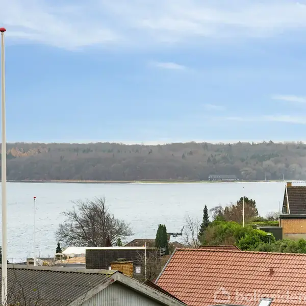 En flagstang står stille i en have med udsigt over en rolig sø omgivet af træer og huse med røde tage på en klar dag.