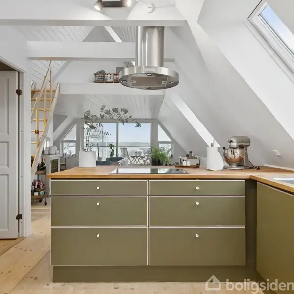 Et køkken med grønne skabe på en ø, hvor der står planter og køkkenredskaber. Et stort ovenlysvindue lyser rummet op. Bagved ses en stige til en hems.