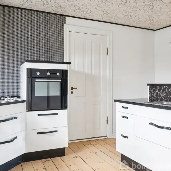 Et moderne køkken med hvide skabe og sorte bordplader dominerer billedet. En ovn er indbygget i skabet. Væggene har mønstrede fliser, og der er en trædør midt i baggrunden. Trægulvet komplementerer køkkenets lyse tema.