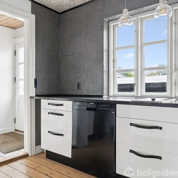 Et moderne køkken med hvide skabe og sort opvaskemaskine. To hængelamper over vindue med udsigt til træer udenfor. Trægulv og tilstødende rum med tæppe og loftslampe.