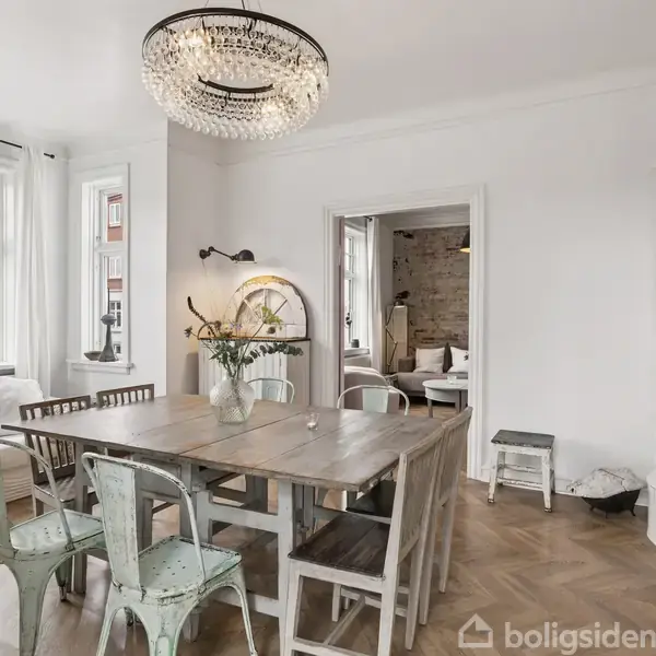 Et moderne køkken med lange bordplader og håndvask, omgivet af hvide vægge og store vinduer. Over bordpladerne hænger en stor lampe. Spisestue ses i baggrunden gennem åben dør.