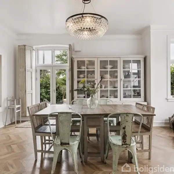 Spisebord omgivet af stole står i et lyst rum med trægulv. En glaslampe hænger over bordet. Bagved er et skab med glaslåger. Store vinduer viser grønne omgivelser udenfor.