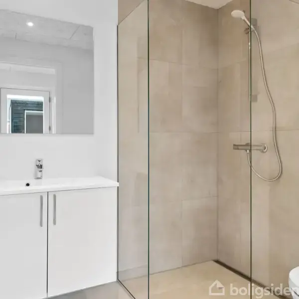 Et moderne badeværelse med en hvid håndvask og skab, et stort vægspejl, og en glasdør til en bruser i et lyst bruseområde med beige fliser.