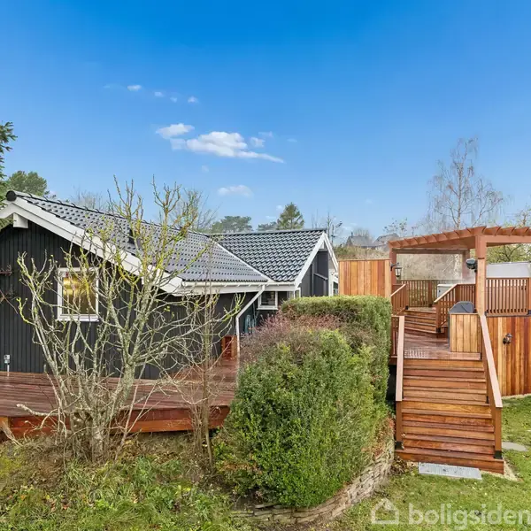Træhus med sort facade og gråt tag står i en have med en stor terrasse. En trappe fører til en hævet, overdækket træstruktur. Baggrunden viser træer og en blå himmel.