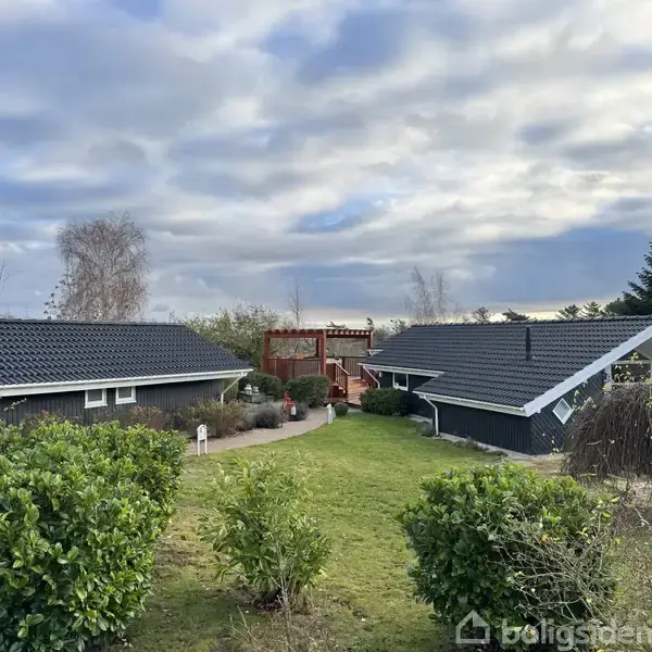 To sorte huse med hvide kanter står i en have med grønne buske og plæne. Mellem husene ses en rød port. Himlen er skyet.