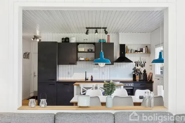 Sort køkken udstyret med moderne apparater og blå lamper over spisebordet; hvidt loft og vægge skaber et lyst og indbydende miljø.