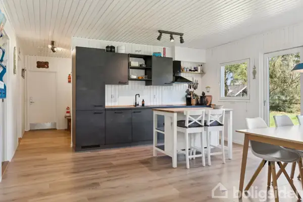 Køkken med sorte skabe står stationært. Hvide vægge omgiver rummet, der fører til en lys spiseplads med træmøbler og vinduer, der viser en grøn have.