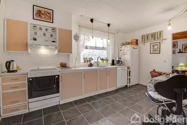 Et køkken med lyserøde skabe, komfur, og ovn på en flisegulv. Køkkenet har en vask ved et vindue med gardiner, omgivet af diverse køkkenudstyr. Væggene er dekoreret med billeder.